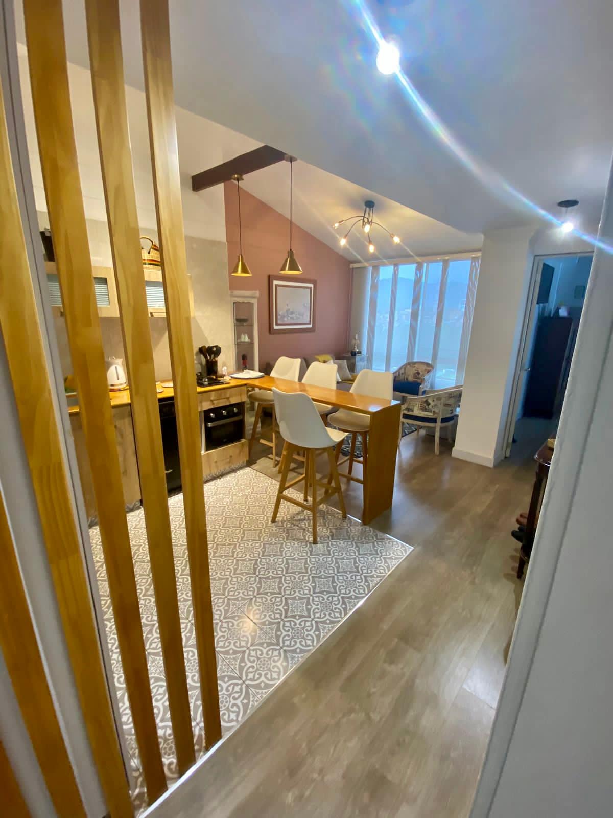The kitchen area is visible, featuring a modern design with wood accents and bar stools. A patterned tile floor adds texture, while natural light enters through large windows. The space leads into a dining area that is softly illuminated, indicating a cozy atmosphere.
