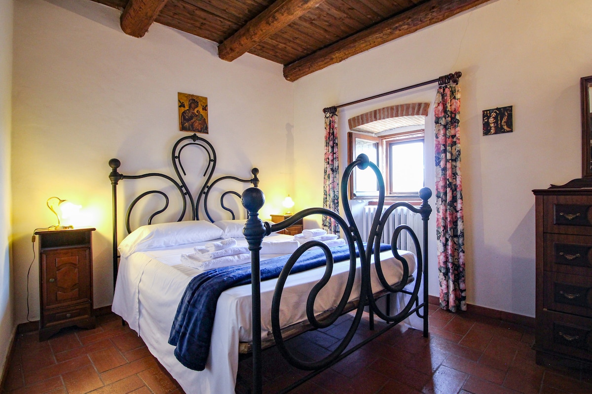 A cozy bedroom features a wrought-iron bed dressed in crisp white linens. Two bedside tables with lamps flank the bed, providing soft lighting. Floral curtains frame the window, which offers a view of the surrounding area. Exposed wooden beams add character to the ceiling.