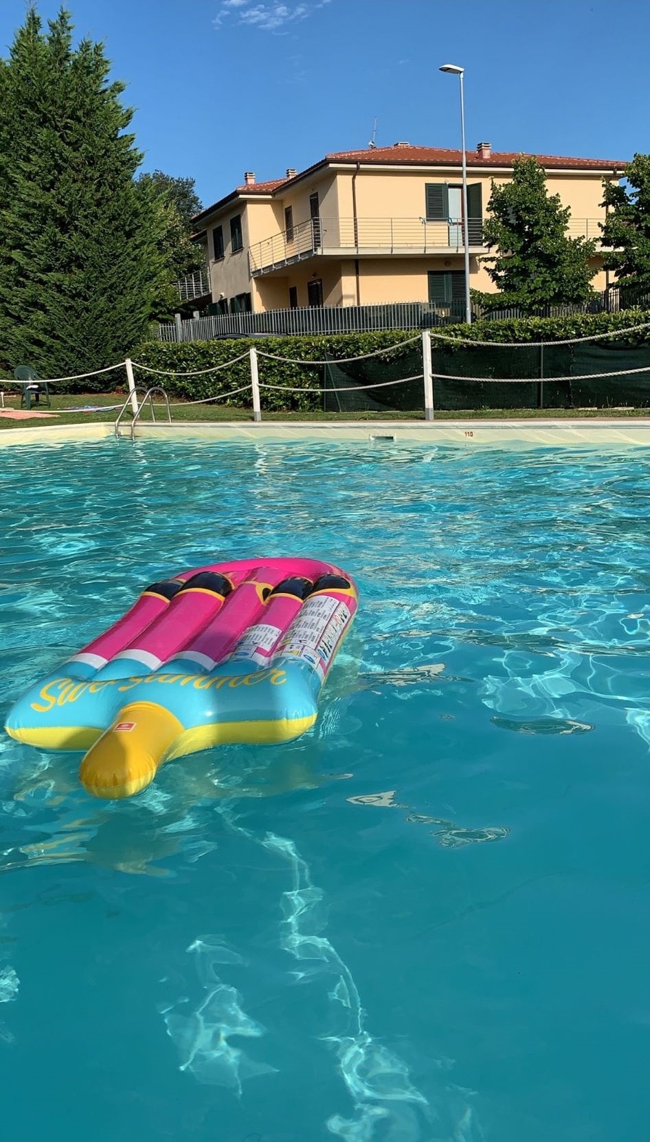 A vibrant inflatable float rests on the surface of a clear blue swimming pool, surrounded by a landscaped area with greenery. In the background, a two-story residential building is visible, framed by trees and a fence.