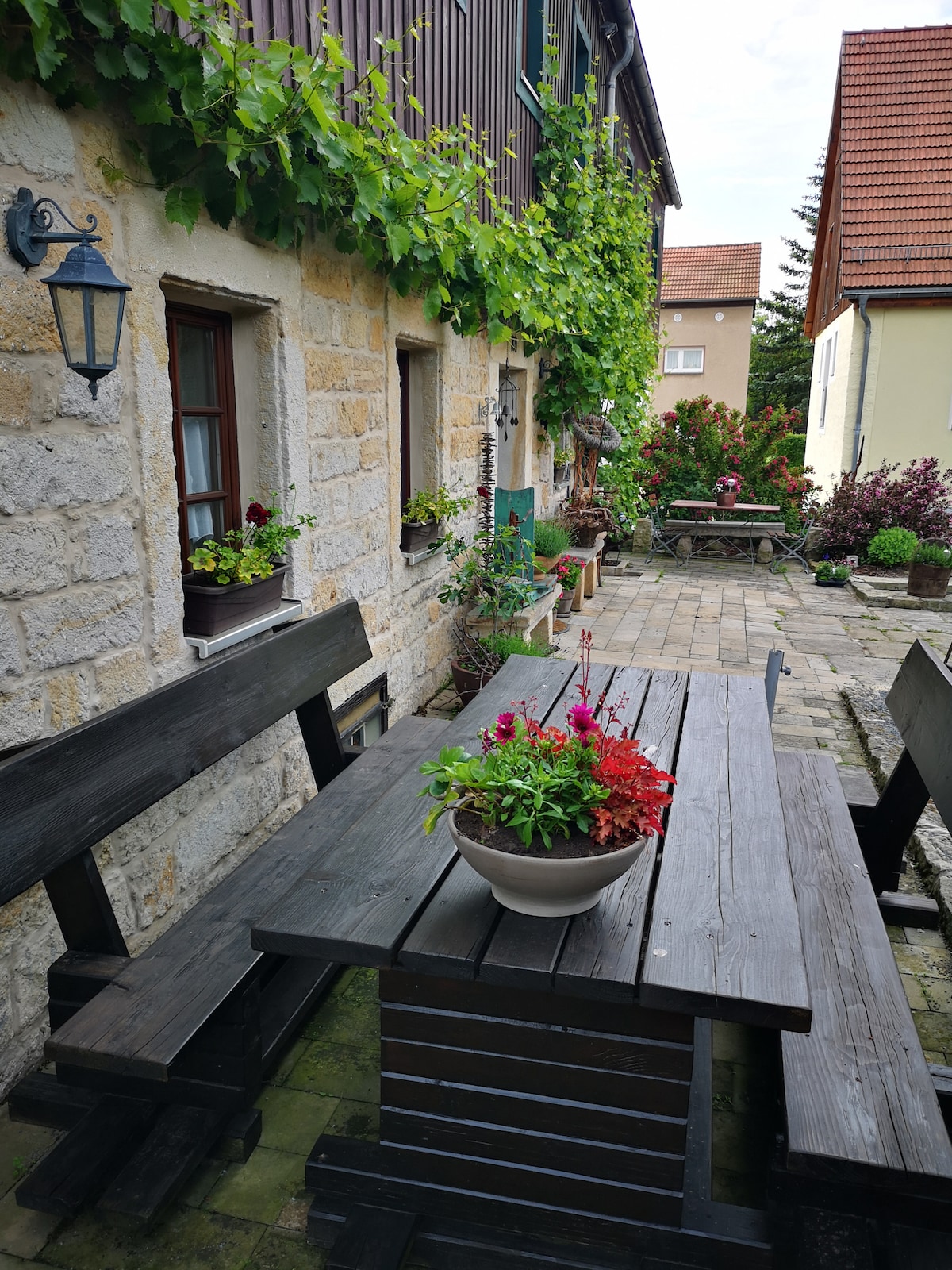 A rustic outdoor dining area features a large wooden table surrounded by benches, adorned with potted flowers. The stone wall of the building, partially covered in greenery, provides a charming backdrop. Additional plants and seating can be seen in the distance, enhancing the inviting atmosphere.