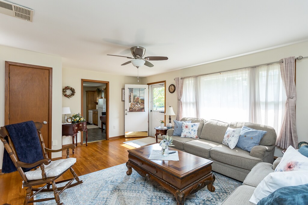 A spacious living room features natural light filtering through large windows. Comfortable seating options include a sectional sofa with decorative pillows and a rocking chair. A wooden coffee table sits in the center, surrounded by a soft rug. The entrance to the kitchen is visible in the background.