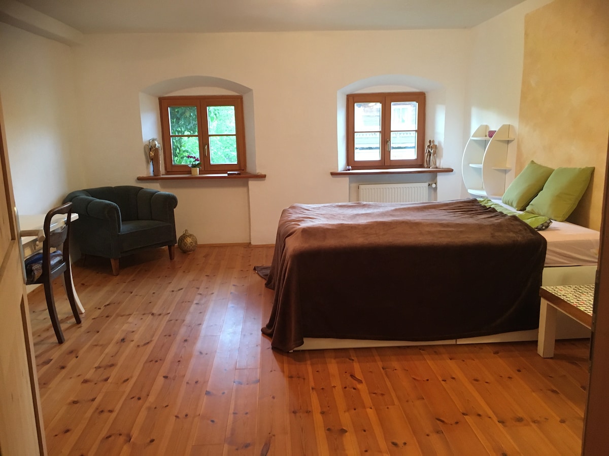 A spacious room features a large double bed with neutral bedding and green accent pillows. Natural light filters through two small windows, illuminating the solid wood floor. A cozy armchair sits in one corner, complemented by a minimalist shelf and a softly painted wall.