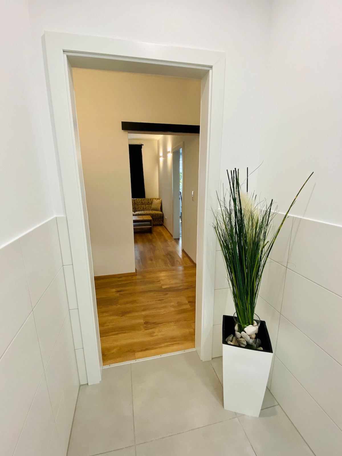 A well-lit hallway is visible, featuring clean, white walls and tiled flooring. A tall, minimalist planter with greenery adds a touch of nature. The space opens into a cozy seating area, framed by an inviting doorway, showcasing wooden flooring.