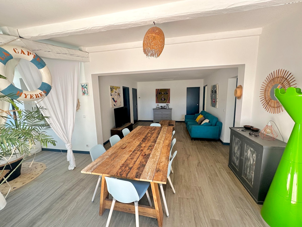 A spacious dining area features a long, rustic wooden table surrounded by light blue chairs. A comfortable teal sofa is positioned nearby. The bright room is adorned with nautical decor and soft lighting, creating an inviting space for gatherings.