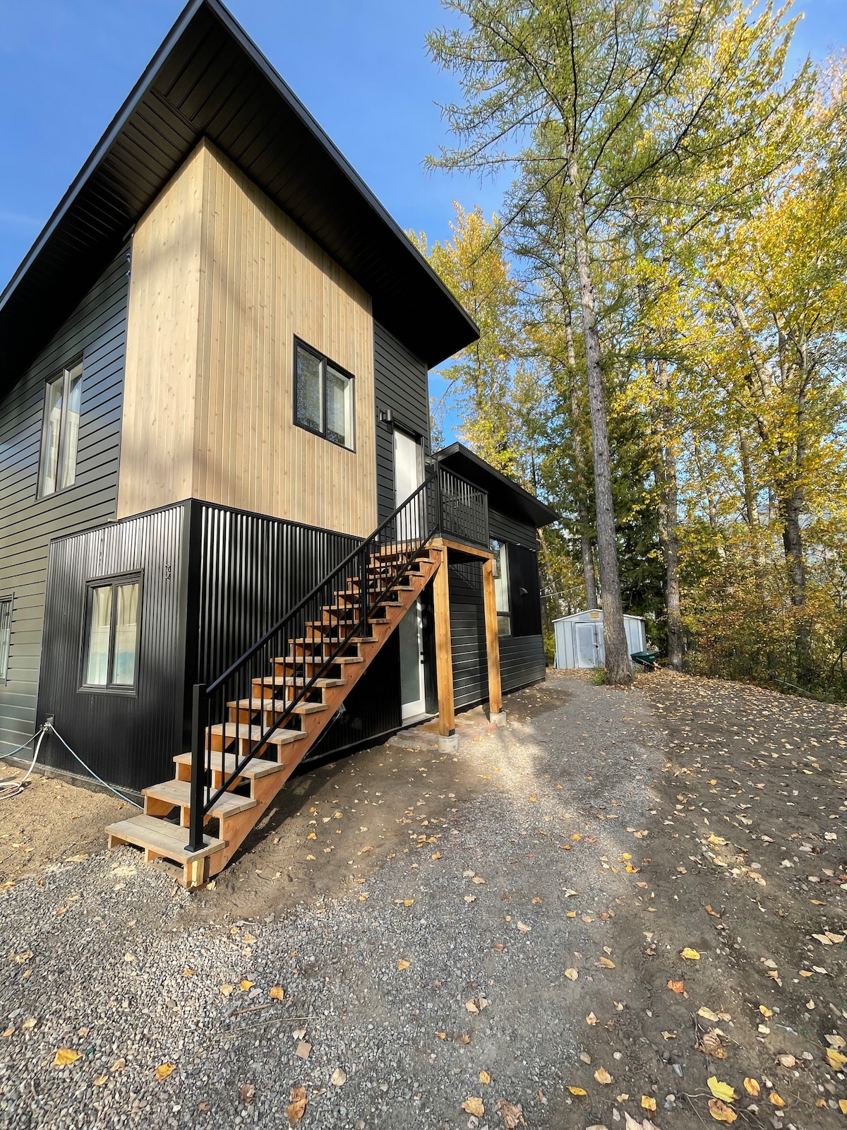 An exterior view of a modern two-story home is showcased, featuring a combination of wood and dark siding. A wooden staircase leads to the entrance, surrounded by a gravel path and nearby trees displaying autumn foliage.