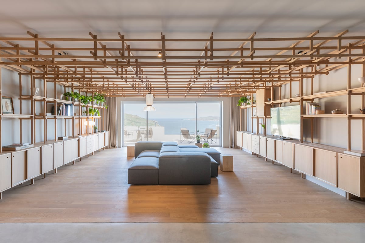 A spacious living area features a minimalist design with wooden shelving and a large sectional sofa. Large glass doors open to reveal scenic views of the ocean and outdoor seating. Natural light fills the space, highlighting the clean lines and open layout.