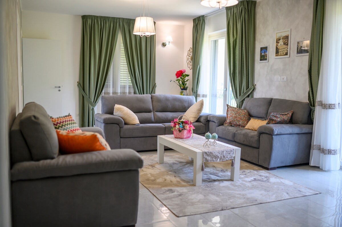 A spacious common area is highlighted by two comfortable sofas, arranged around a central coffee table. Soft lighting fixtures illuminate the room, while green drapes frame the windows, allowing natural light to enter. Decorative elements, including cushions and wall art, add warmth to the space.