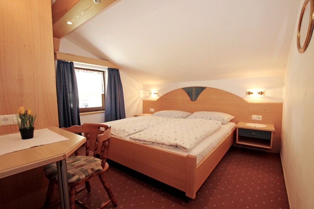 A cozy sleeping area features a spacious bed with light bedding. A small table and chair are positioned nearby, providing a space for relaxation. Dark curtains frame the window, and soft lighting enhances the inviting atmosphere.