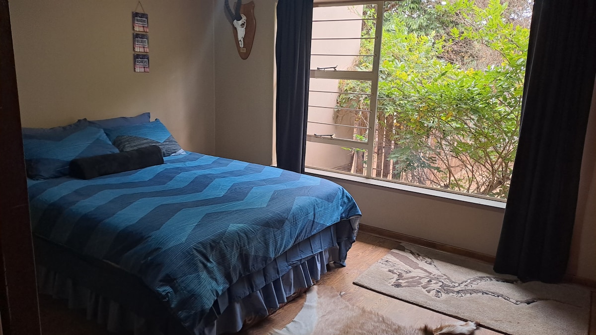 A spacious sleeping area features a plush blue bedspread with matching pillows. Sunlight enters through a large window, illuminating the room and showcasing greenery outside. A stylish area rug is placed on the wooden flooring, contributing to a cozy ambiance.