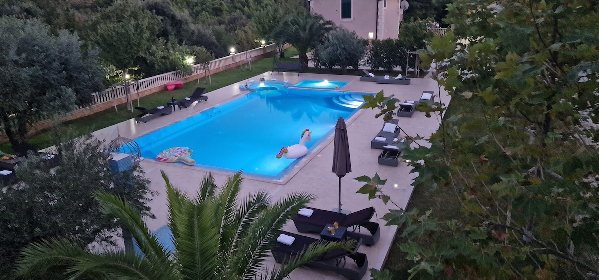 An expansive swimming pool is surrounded by landscaped gardens, featuring comfortable lounge chairs and a poolside umbrella. Colorful inflatables float on the water, while palm trees and greenery enhance the overall tranquil atmosphere of the outdoor space.