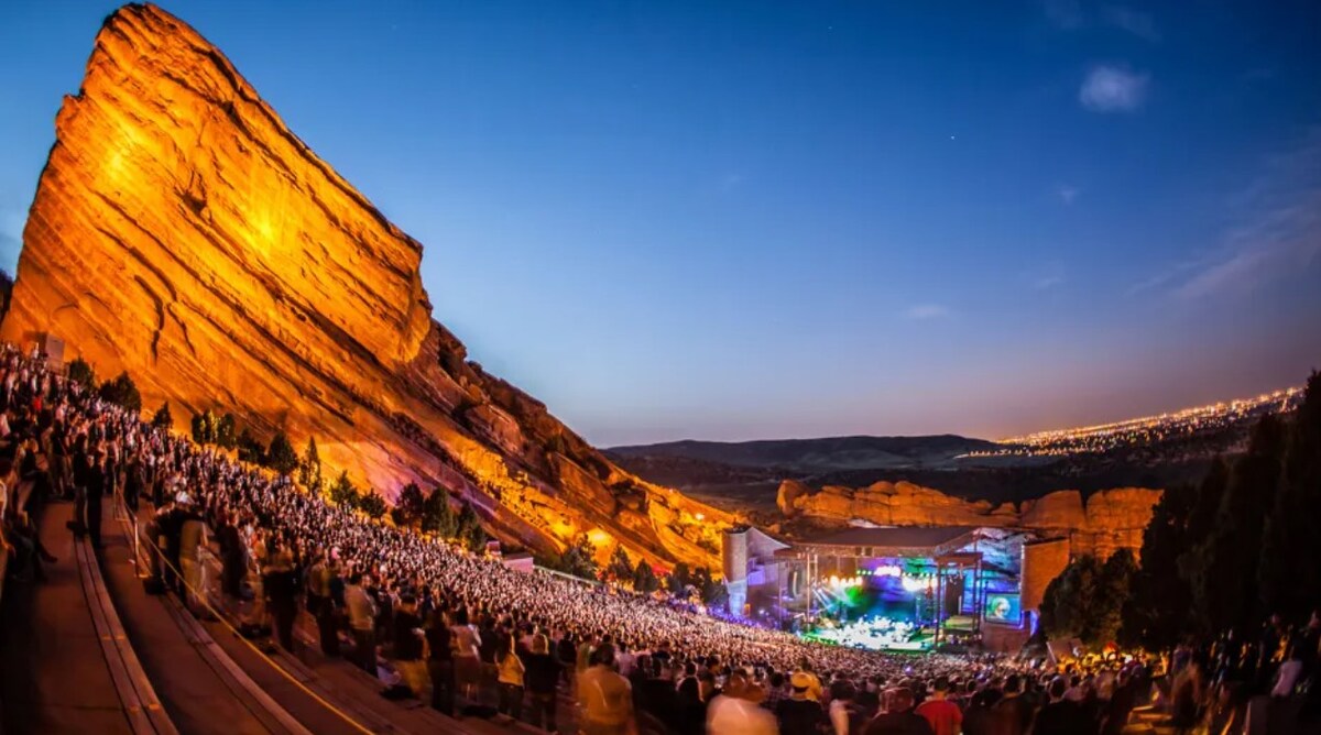 A large outdoor amphitheater is shown, nestled between towering red rock formations that are illuminated by soft golden light. The audience is gathered, enjoying a concert with a vibrant stage set against the extensive backdrop of the surrounding landscape.