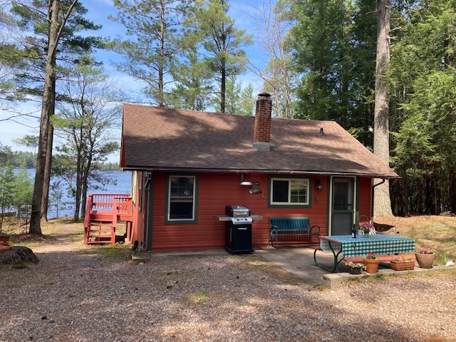 A charming cottage is situated among tall trees, with a view of the serene lake in the background. A deck and outdoor seating area are accessible, accompanied by a grill and table set with colorful flower pots, creating a welcoming space for relaxation.