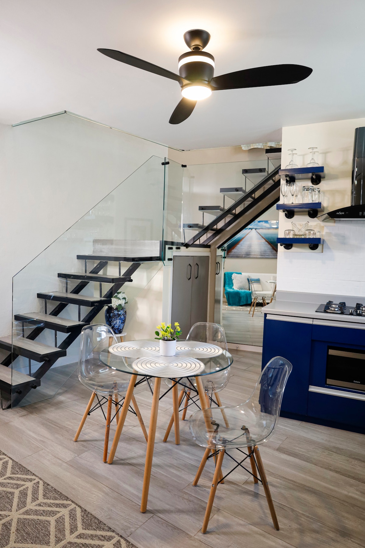 A modern dining area features a round glass table surrounded by transparent chairs. A ceiling fan provides ventilation. A staircase with glass railing leads to the upper level. The kitchen, equipped with sleek cabinetry and modern appliances, is visible in the background.