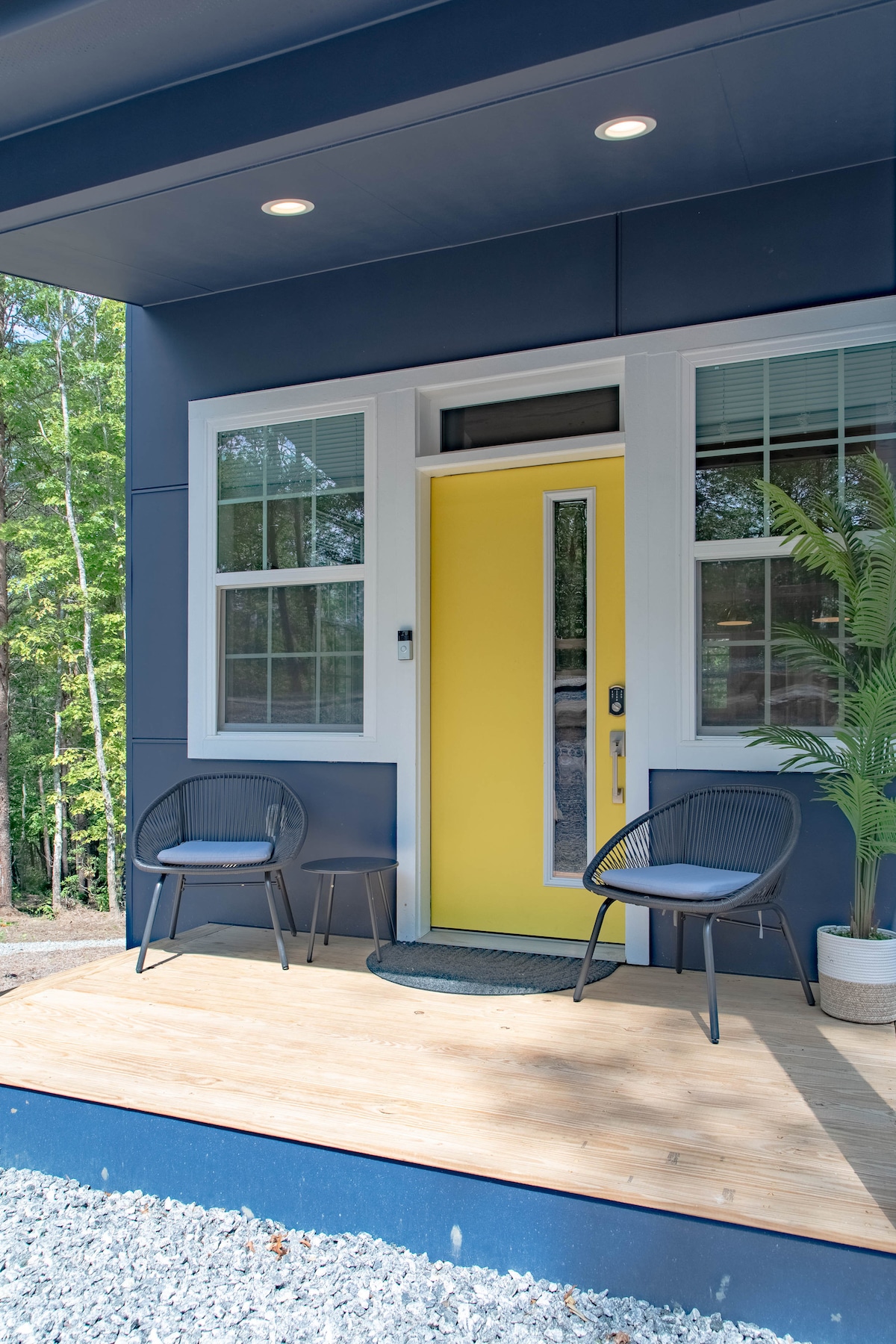 A welcoming entrance is presented with a bright yellow front door, flanked by two black chairs on a wooden porch. Sunlight filters through the windows, which are adorned with white trim, revealing a glimpse of surrounding greenery.