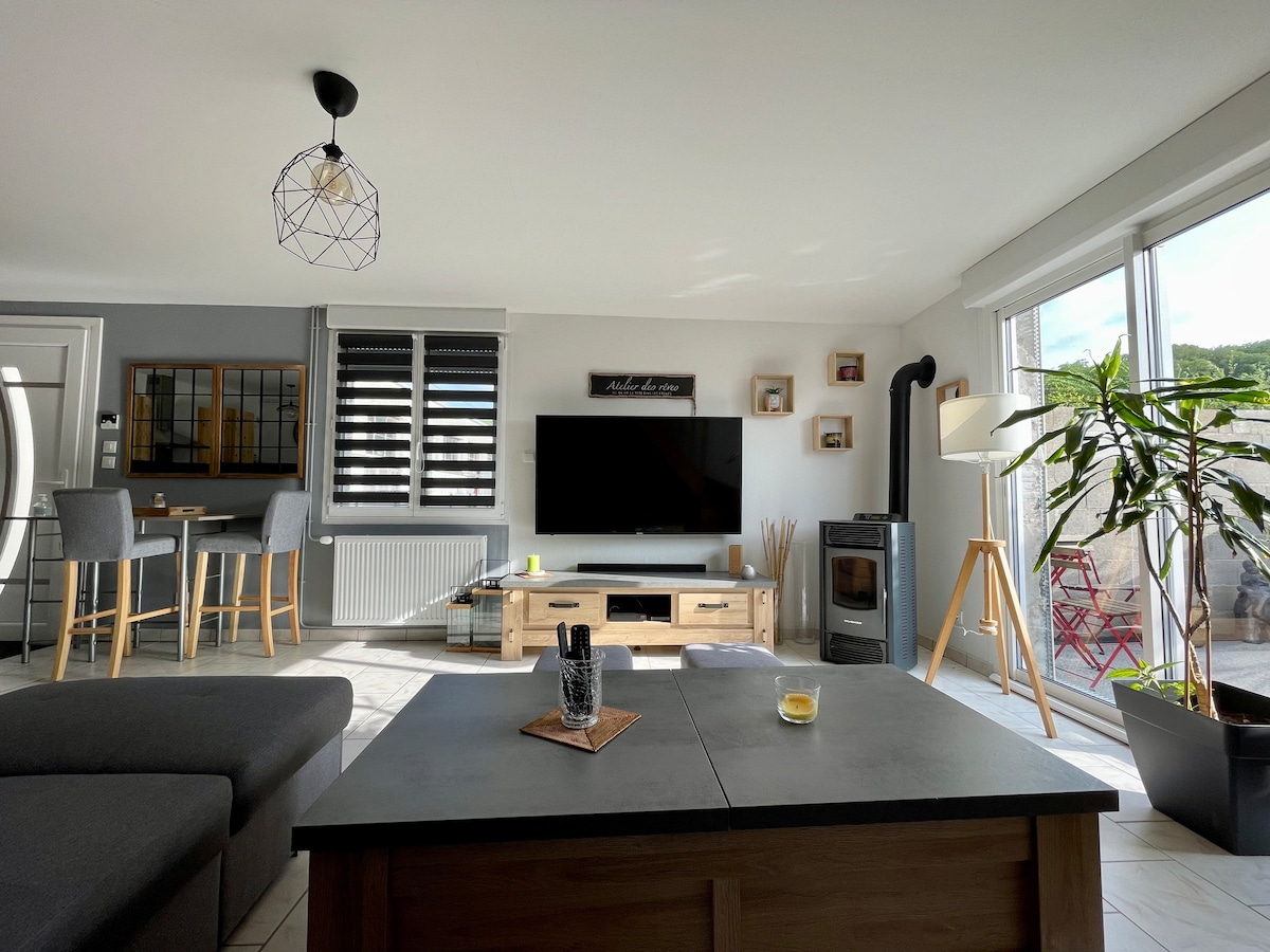 A spacious living area features a gray sofa and a modern coffee table at its center. A large television is mounted on the wall opposite the seating. Natural light floods in through large windows, illuminating a dining table and chairs in the background. A decorative plant is also visible.