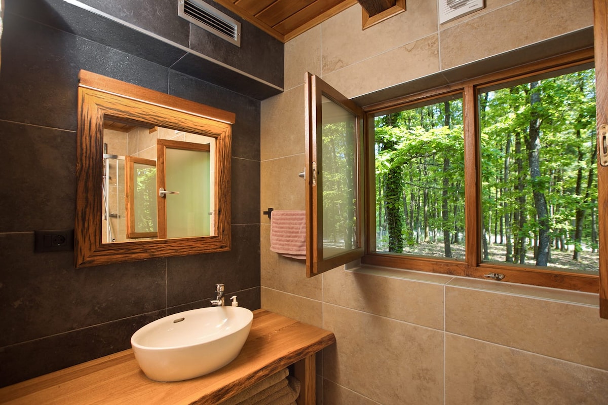 A modern bathroom features a sleek wooden countertop with a white basin. Natural light is provided by a window that opens to a forest view, framed by two wooden panes. A mirror with a wooden frame hangs above the sink, complementing the warm tones of the space.
