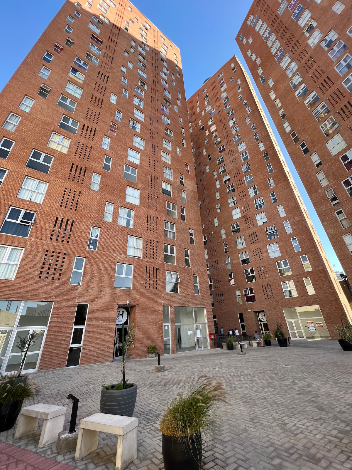A modern multi-story building showcases a brick facade with numerous windows. The entrance features a clock and is surrounded by landscaped planters, adding a touch of greenery to the structure. The blue sky enhances the overall appearance of the building.