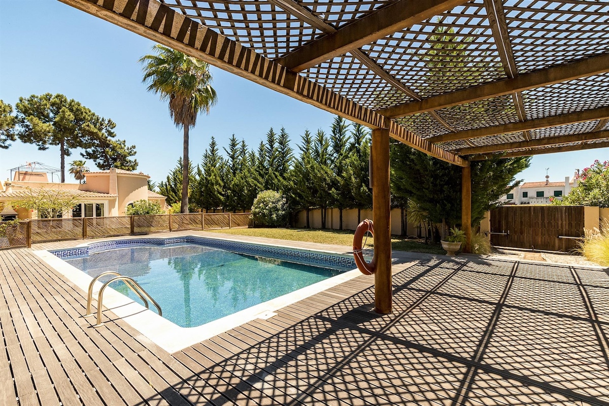A private swimming pool is surrounded by wooden decking, featuring a protective pergola overhead. Lush greenery lines the perimeter, while a palm tree adds a tropical touch. A life buoy is positioned near the pool's edge, enhancing safety for swimmers.