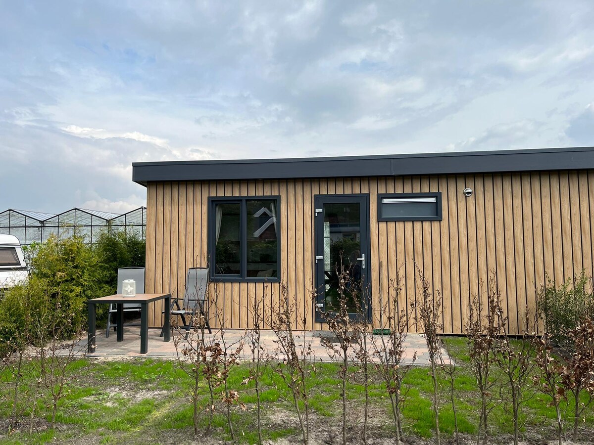 A modern wooden exterior features large windows and a welcoming entrance. A small table and two chairs are positioned on the patio, surrounded by well-maintained grass and low shrubs. The structure is complemented by a clear sky and distant greenery.