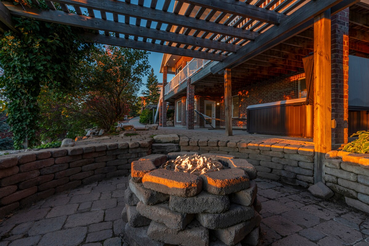 A gas fire pit is surrounded by a circular stone seating area, set on a patio paved with rectangular stones. Wooden beams create a pergola overhead, casting gentle shadows. The evening light enhances the warmth of the scene, highlighted by lush greenery in the background.