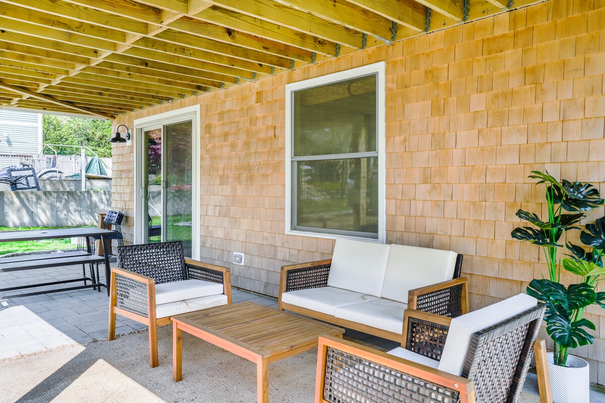 An outdoor seating area is presented with a wooden table and four chairs arranged underneath a covered space. Light-colored walls enhance the inviting ambiance, while a green potted plant adds a touch of nature. Large windows allow for natural light and ventilation.