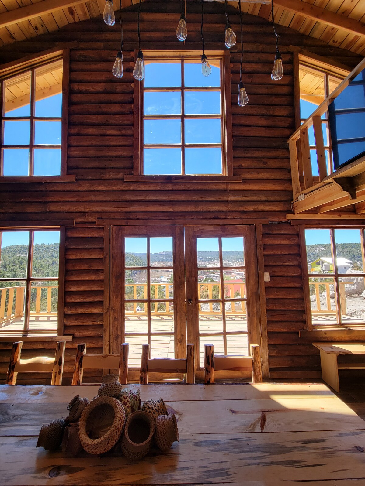 Large windows allow natural light to fill the cabin's interior, illuminating a wooden dining table adorned with woven baskets. The spacious area offers views of the surrounding mountains and the outdoor balcony, enhancing the connection to nature.