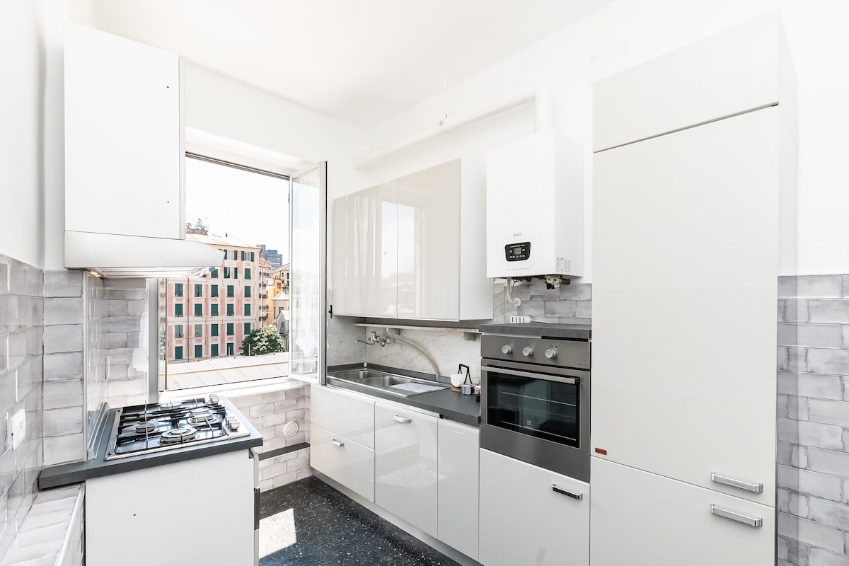 A modern kitchen is presented with white cabinets and sleek surfaces. Large windows allow natural light to fill the space, highlighting the gas stove and oven. The kitchenette includes a sink and ample countertop space, contributing to a functional and welcoming environment.