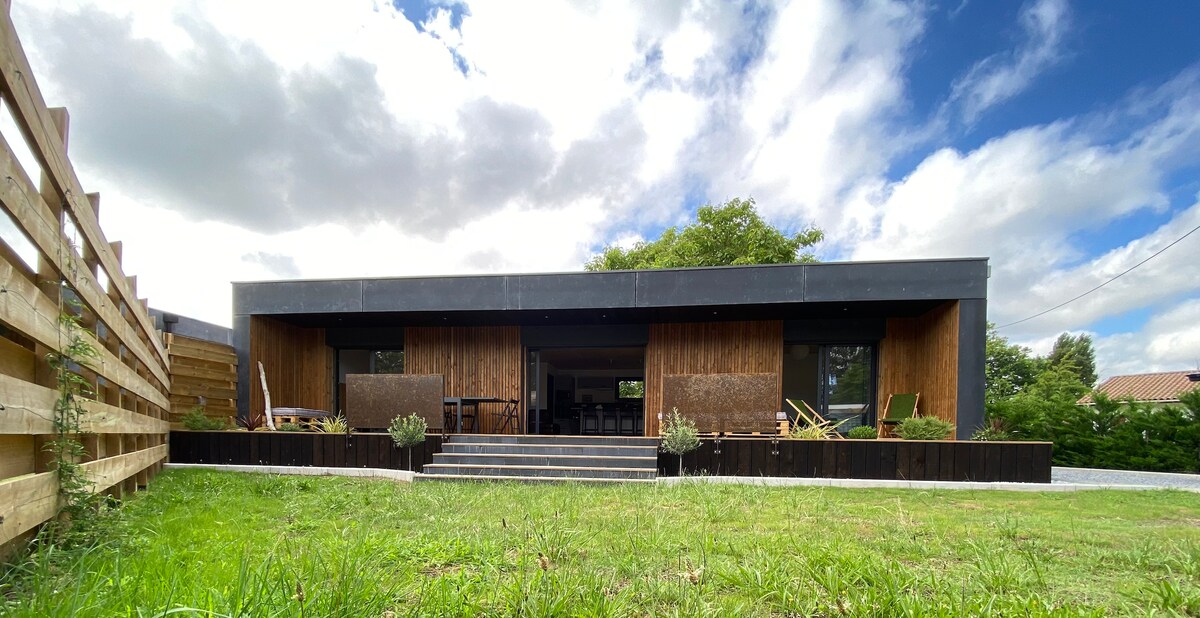 The modern house features a large wooden facade with floor-to-ceiling windows, allowing for natural light. A spacious terrace is present, bordered by greenery, and steps lead to a well-maintained lawn. The structure is complemented by a clear blue sky with scattered clouds.