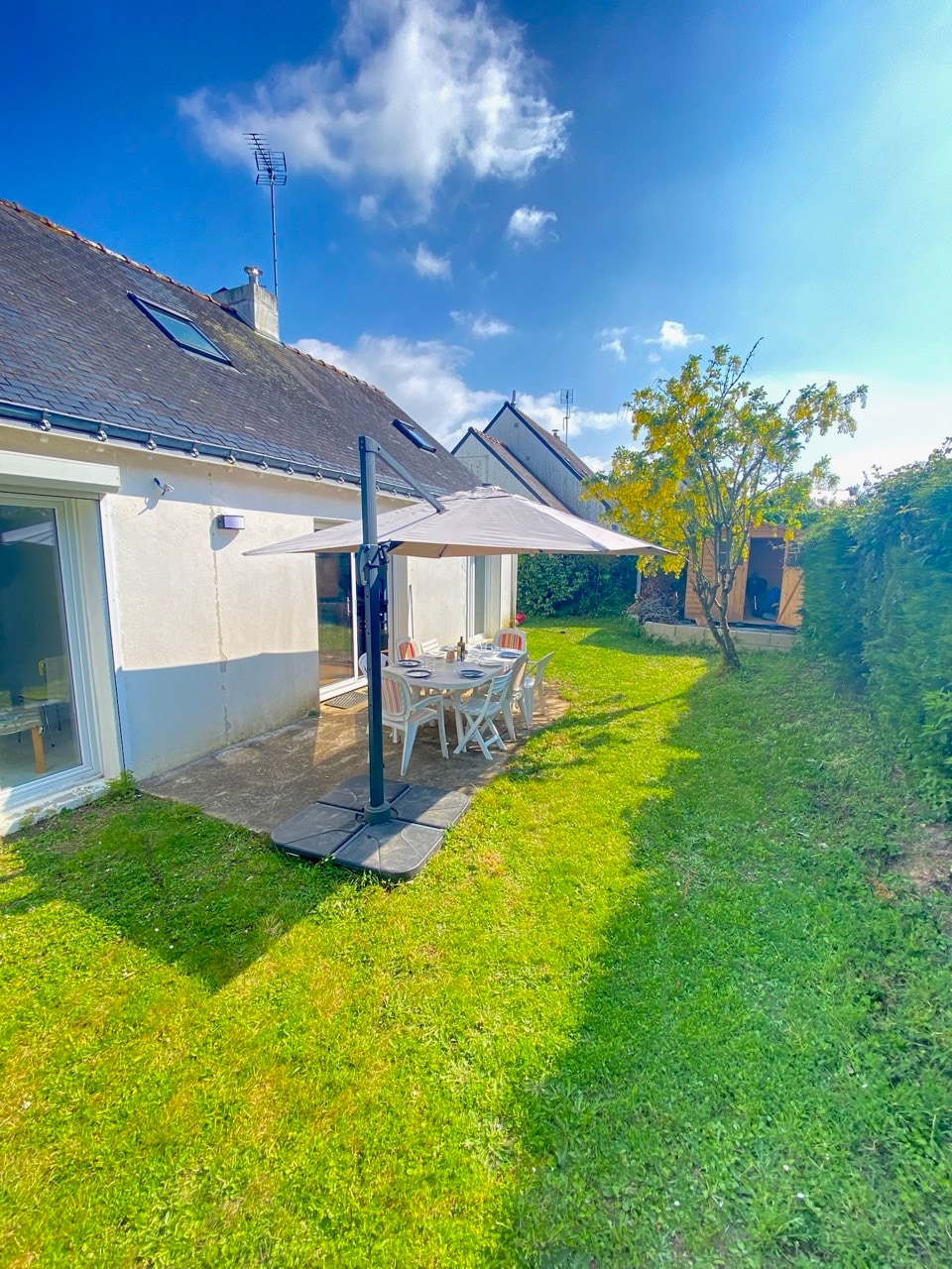 A sunny garden area features a table set for outdoor dining under a white umbrella. The well-maintained lawn is bordered by green shrubs, and the house's exterior is illuminated by natural light, with large windows reflecting the blue sky.