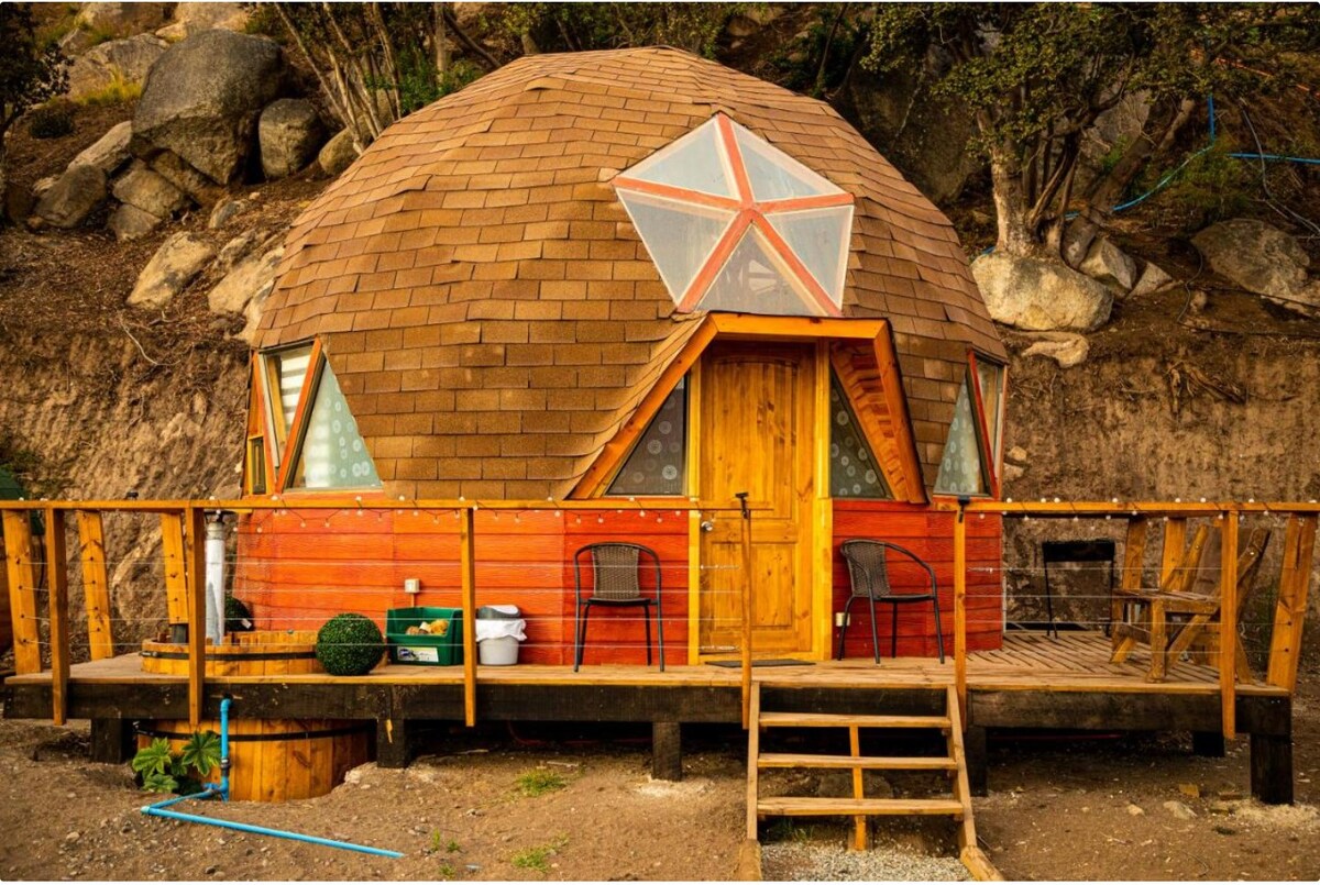 A round dome structure is surrounded by nature, featuring a wooden deck with railings and steps leading to the entrance. Large triangular and circular windows offer natural light, while two chairs sit outside, inviting relaxation. The earthy tones of the exterior blend harmoniously with the landscape.