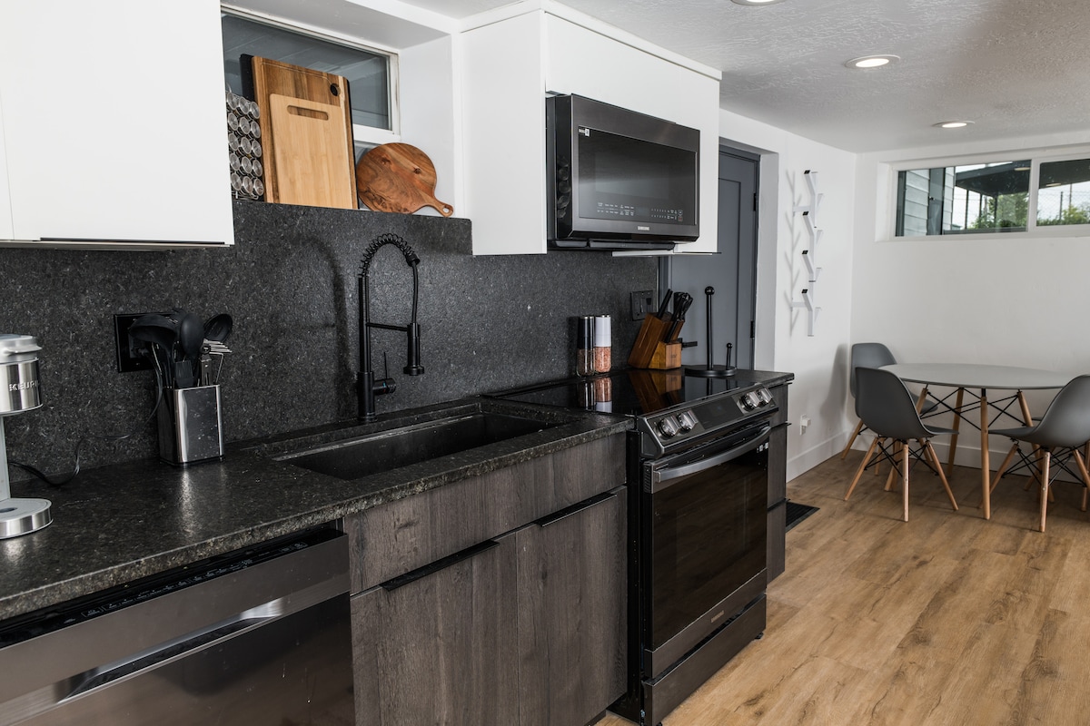 A modern kitchen is equipped with stainless steel appliances, including a sleek stove and microwave. Dark countertops are complemented by light cabinetry, while a dining table with a few chairs is visible in the background. Natural light enters through a nearby window.