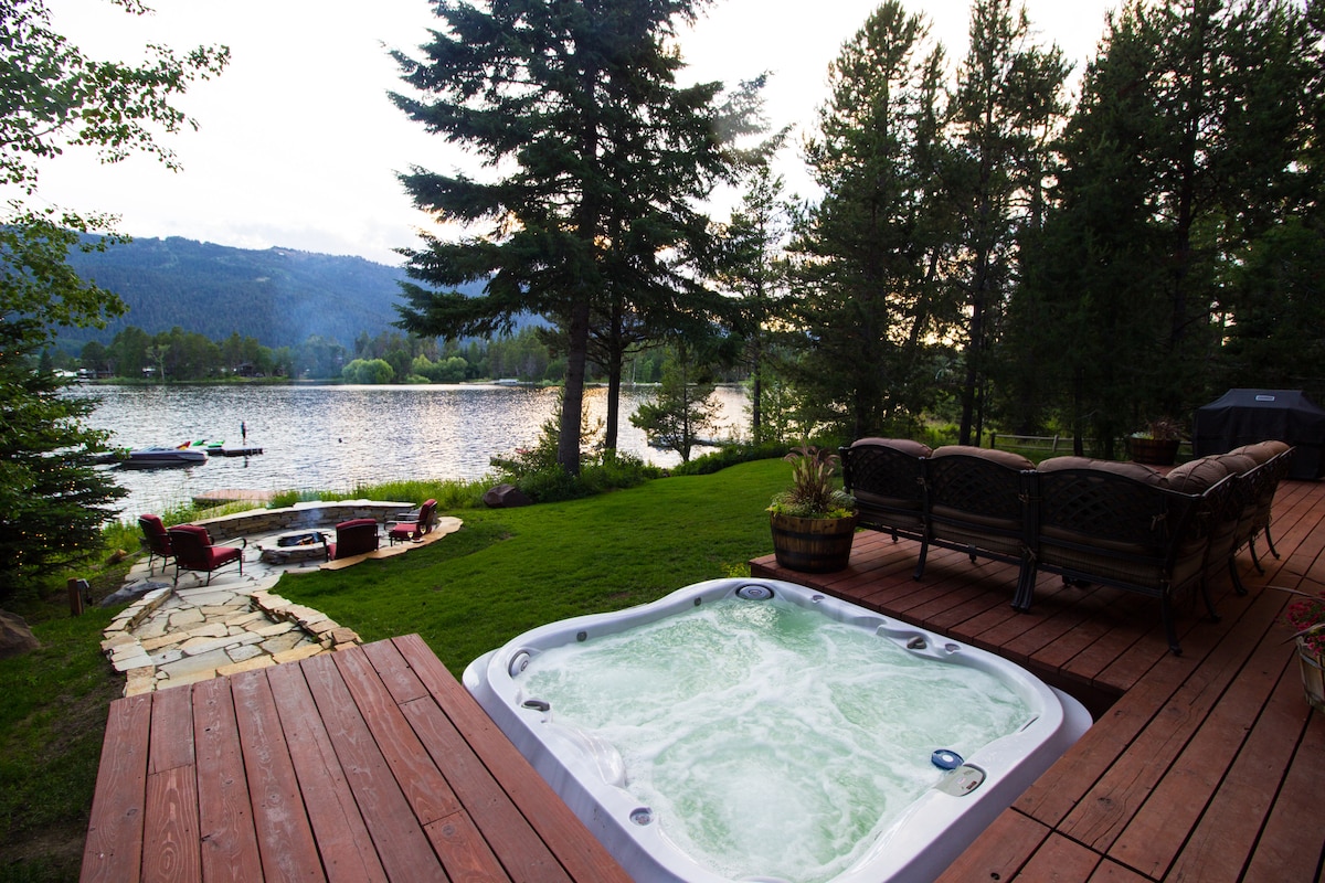 A hot tub is situated on a wooden deck overlooking a serene lake, surrounded by trees and greenery. Comfortable patio seating is arranged nearby, with a view of the water and distant mountains. A peaceful atmosphere is created by the natural setting.
