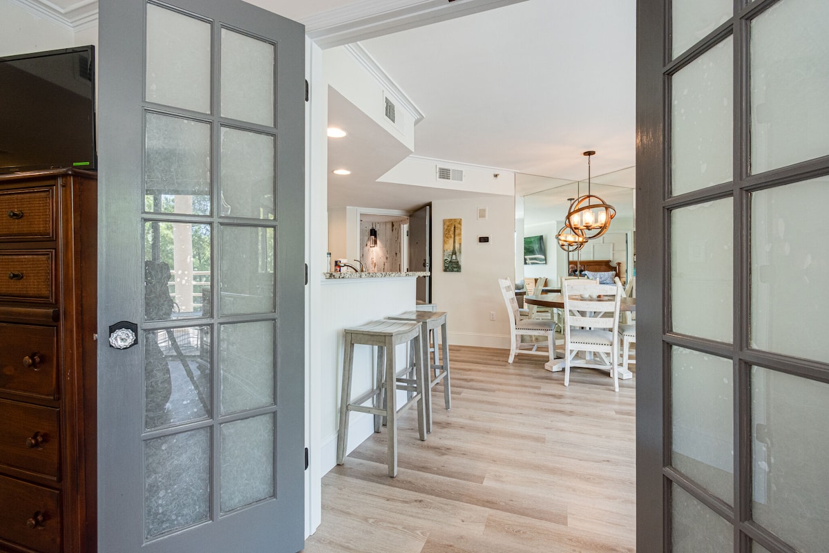 An entrance view showcases an open floor plan leading into the kitchen and dining area. Light wood flooring complements soft wall colors. Glass-paneled double doors provide a glimpse of the dining set, illuminated by a modern pendant light.