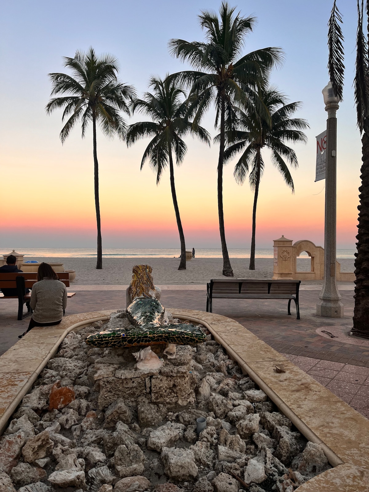 A serene beach scene is captured during twilight, featuring palm trees reaching towards a softly lit sky. A decorative fountain made of natural stones is positioned in the foreground, with gentle waves lapping at the sun-kissed shoreline in the background.