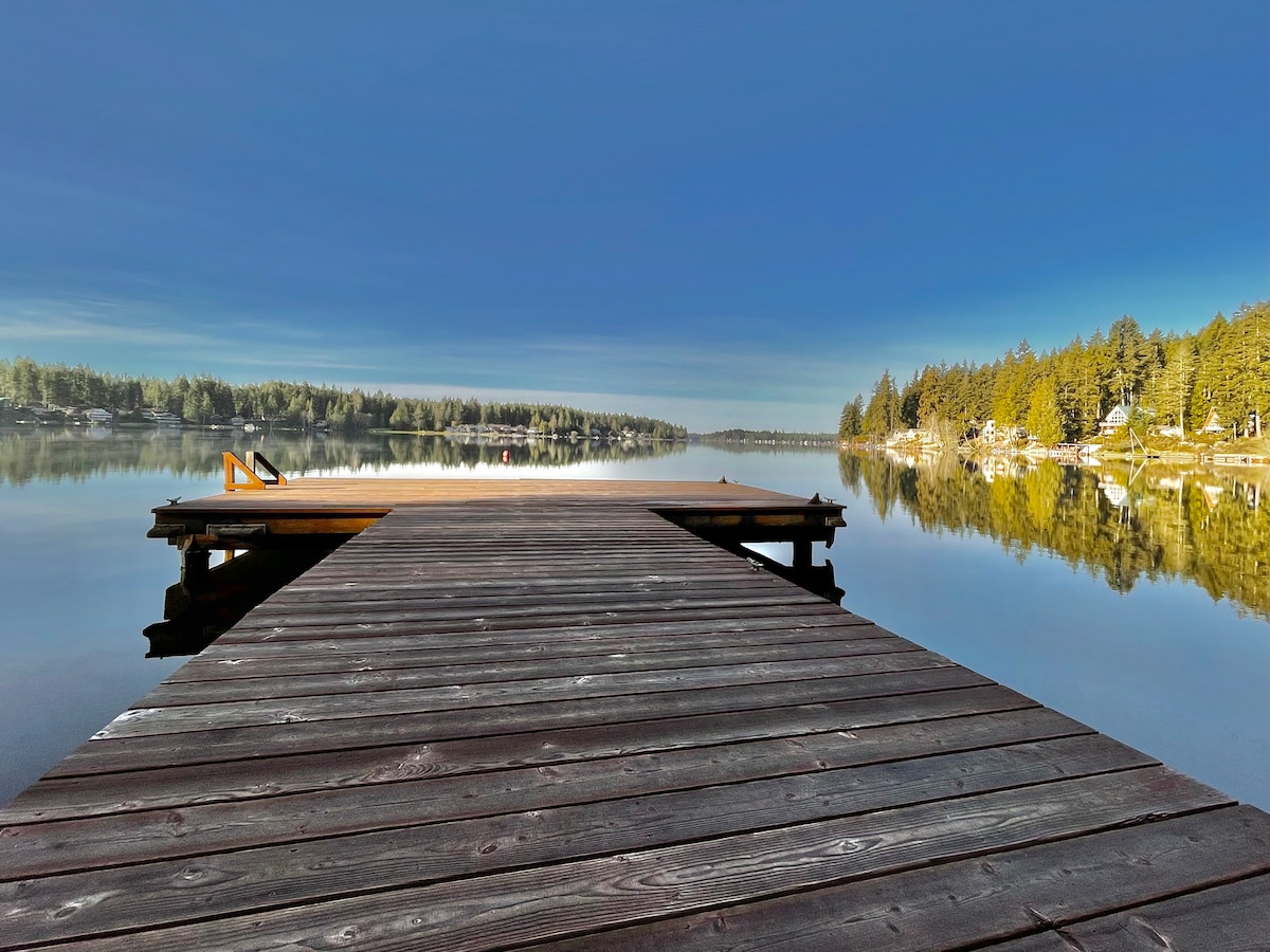 A wooden dock extends over calm waters, reflecting the surrounding trees and sky. A single orange chair sits at the edge of the dock, inviting relaxation. The scene is framed by gentle hills and a serene landscape of pine trees beyond the water.