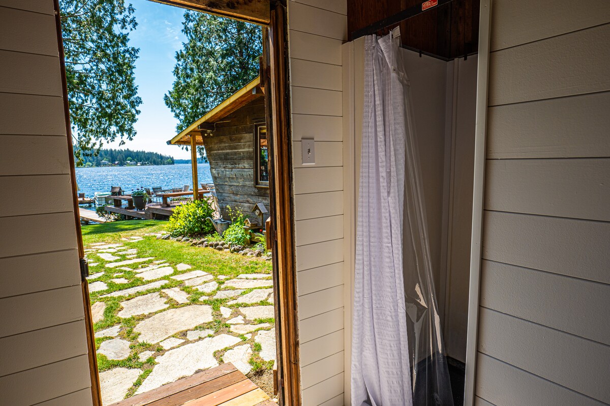 The image depicts an open door leading to a stone pathway that connects to a lakefront area. Sunlight brightens the space, highlighting the natural surroundings and a glimpse of the shore with wooden seating along the water.