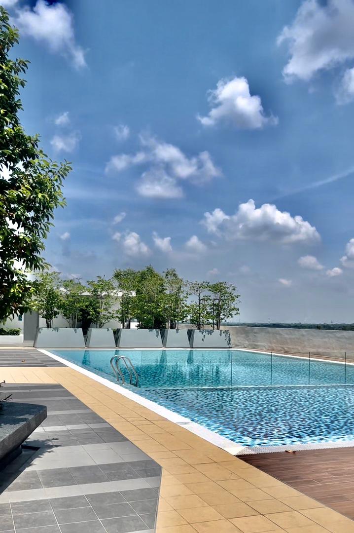 An outdoor swimming pool features clear blue water, bordered by tiled decking and surrounded by greenery. The bright sky displays scattered clouds, enhancing the tranquil setting. A path leads from the pool area toward the distant horizon, offering a picturesque view of the landscape.