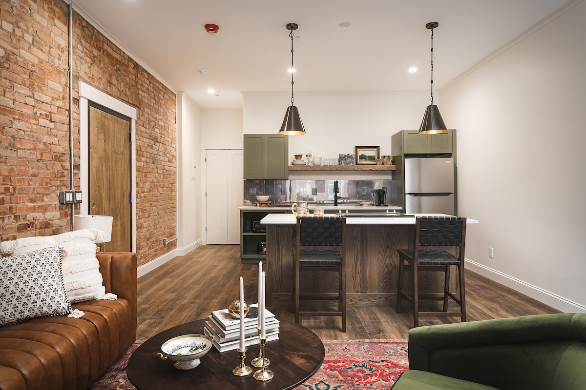 An open living space features exposed brick walls and stylish furnishings. A comfortable brown sofa and green armchair surround a dark circular table, which displays decorative items. The kitchenette is equipped with modern appliances and features bar-style seating.