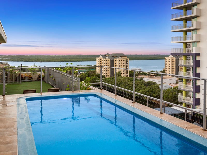 A gleaming rooftop pool reflects the vibrant hues of the sunset, with panoramic views of the ocean and cityscape. Comfortable lounge chairs are positioned beside the pool, inviting relaxation in the serene atmosphere. Green expanses are visible in the distance, framed by architectural structures.