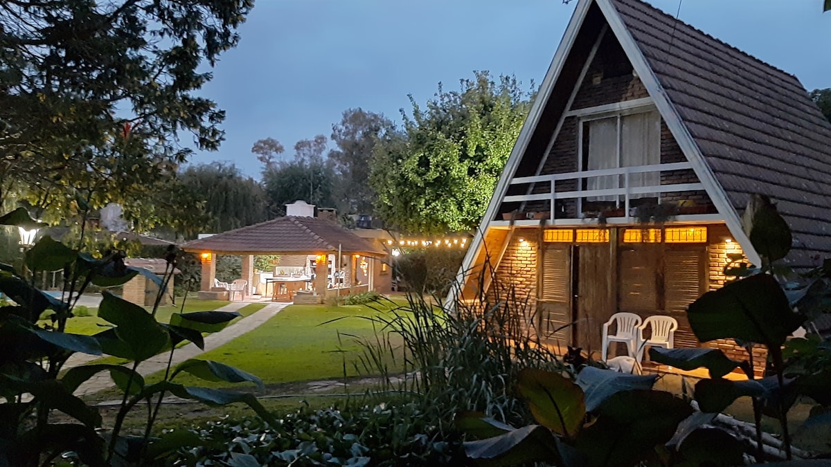 A charming cabin and a spacious quincho are viewed from a garden filled with greenery. String lights illuminate the outdoor area. The cabin features a balcony, while cozy seating arrangements are visible near the quincho, surrounded by trees and a well-kept lawn.