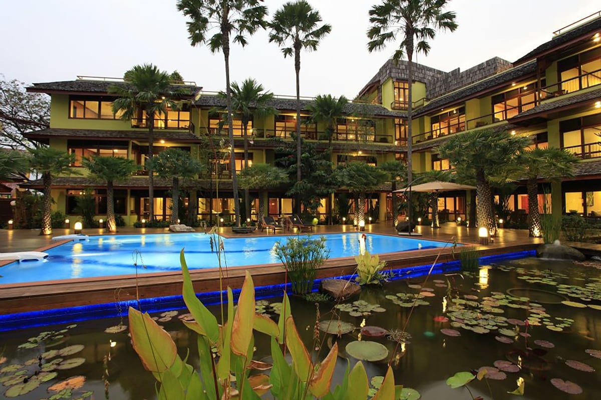 An outdoor swimming pool is framed by lush greenery and palm trees, with tranquil water features and lotus plants in the foreground. The multi-level building surrounds the pool, showcasing large windows that reflect the natural surroundings, creating a peaceful ambiance.
