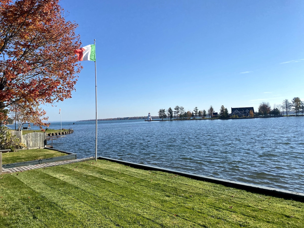 A scenic view of the lake is captured, featuring calm waters under a clear blue sky. A grassy area is framed by a neatly maintained lawn. A pole with a flag stands on the left, while a lighthouse is visible in the distance, accompanied by a cluster of trees.