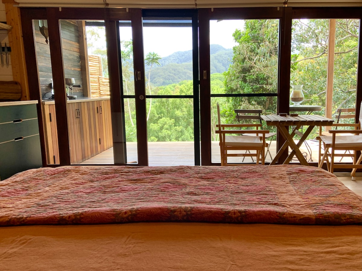A spacious bed draped with a textured blanket faces large glass doors that open to a view of lush greenery and distant mountain ranges. A wooden dining table and chairs sit on the deck, inviting outdoor relaxation.