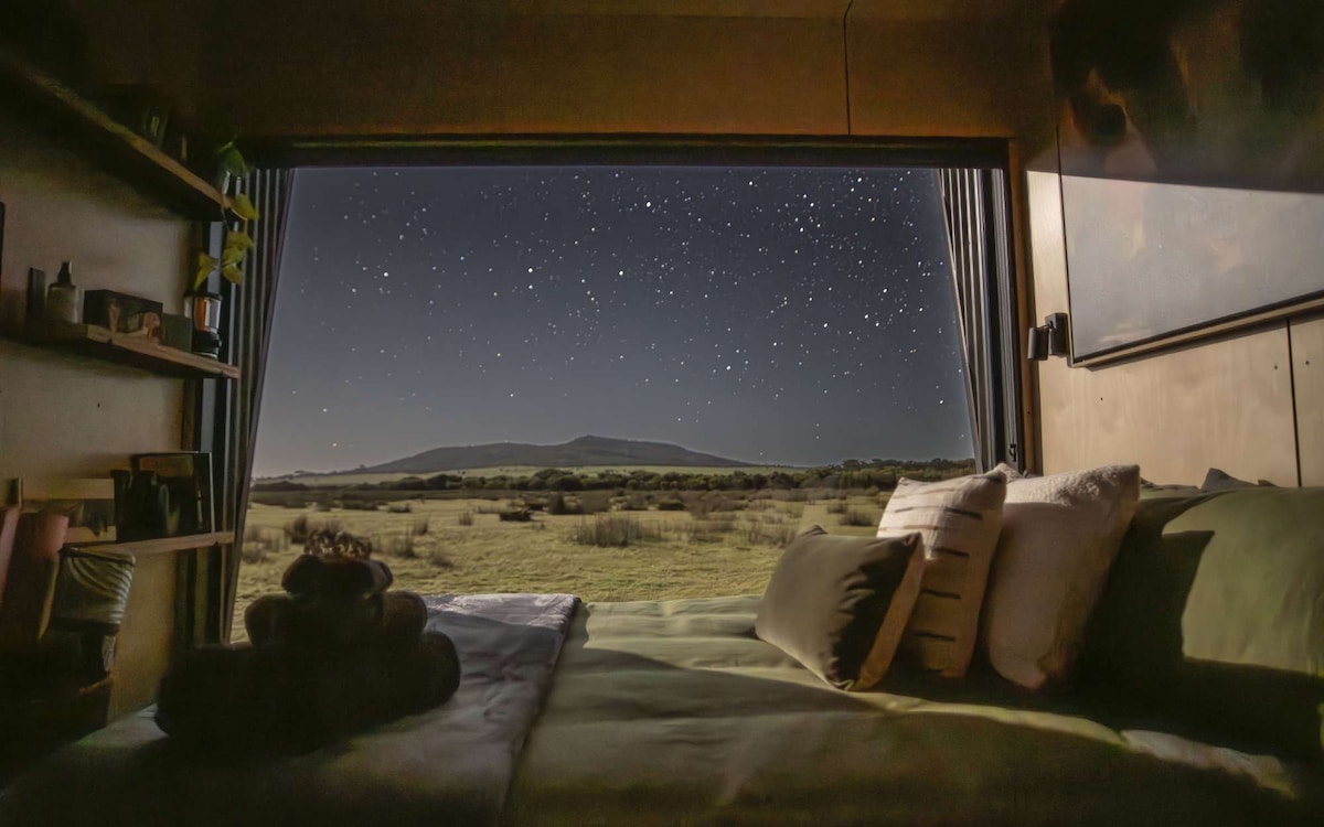 A cozy sleeping area offers a view of a starry night sky through floor-to-ceiling glass doors. A spacious bed is adorned with decorative pillows and blankets. The surrounding landscape features gentle hills, enhancing the tranquil setting.
