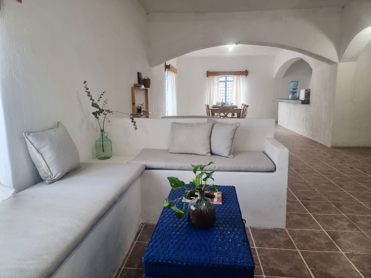 A welcoming seating area features a light-colored sofa with soft cushions. A small blue table holds a vase with greenery. Arched openings lead to a dining area and a kitchen beyond, where natural light filters through sheer curtains.