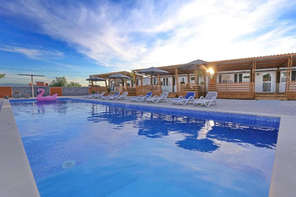 A tranquil swimming pool is bordered by a gravel area, featuring a series of white sun loungers and umbrellas. A pink inflatable float is visible in the water, while a set of wooden structures sits in the background, providing shade and relaxation.