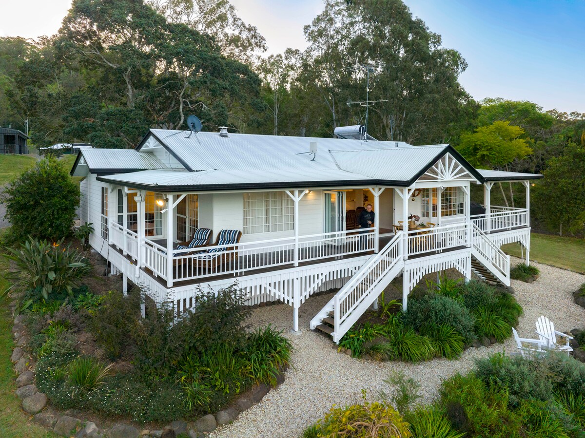 The exterior of a spacious country house is displayed, featuring a wide verandah with seating and large windows. Surrounded by lush landscaping and trees, the home is set against a clear sky, showcasing a warm and welcoming atmosphere.