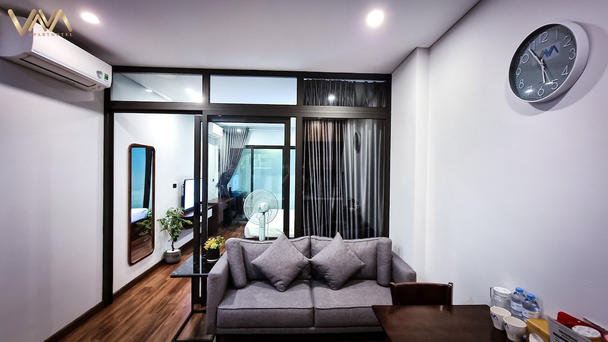 A comfortable seating area features a gray sofa adorned with pillows. A wooden table is arranged nearby, complemented by water bottles. Large glass doors, dressed with sheer curtains, lead to a bedroom area visible in the background. A clock is mounted on the wall above.