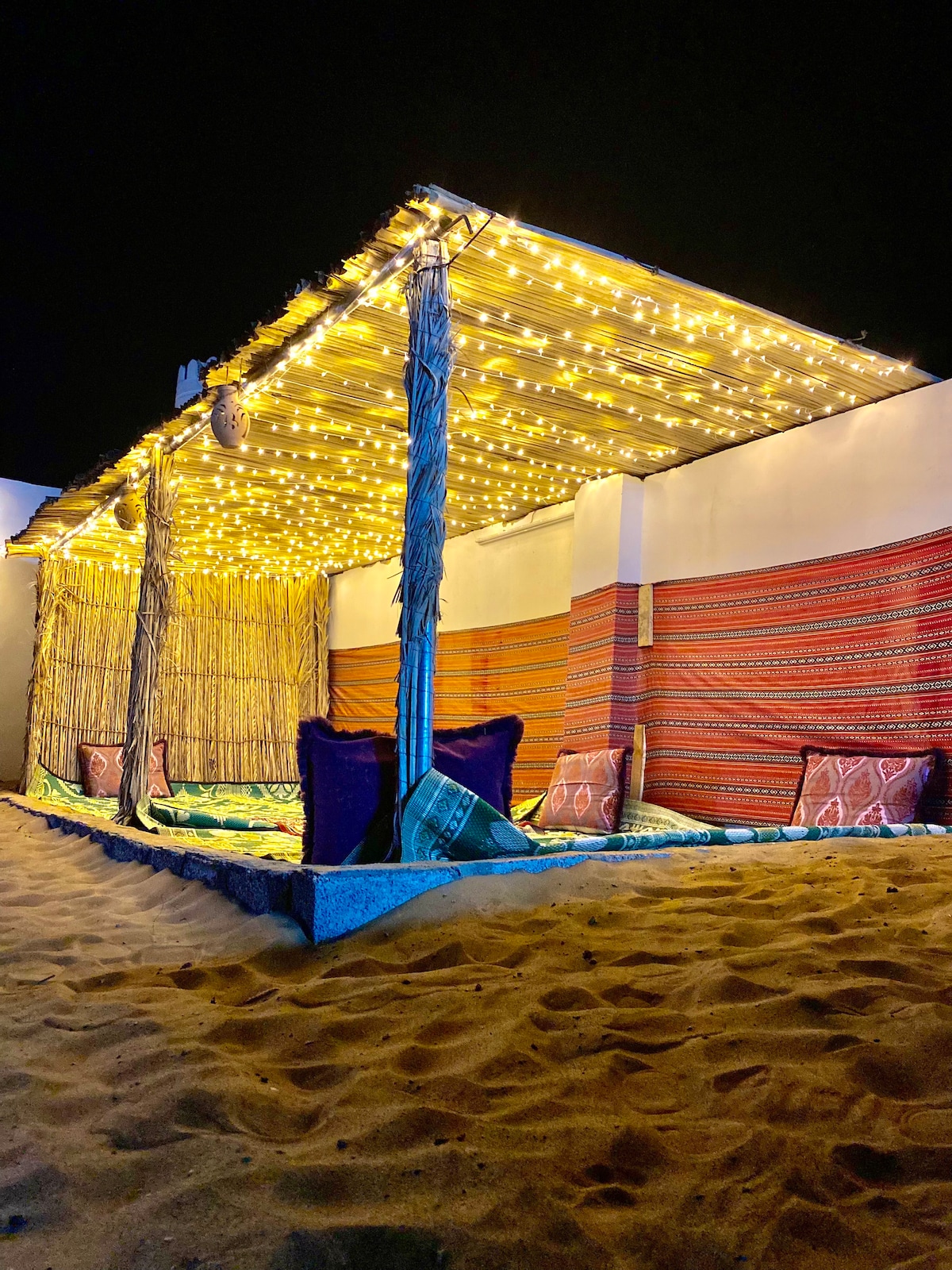 A shaded seating area is adorned with string lights, illuminating the space with a gentle glow. Colorful woven wall hangings and cushions are arranged around a sandy floor, creating a cozy environment for relaxation under the night sky.