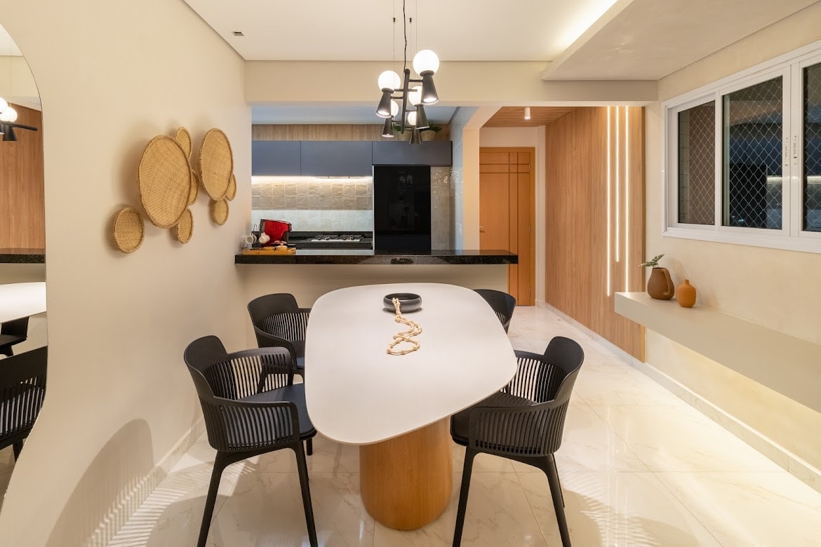 A modern dining area features a sleek oval table surrounded by black chairs. A minimalist wall display of woven decor adds texture, while a well-lit kitchen is visible in the background. Natural light filters through a window, enhancing the space's open and inviting feel.